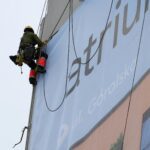 Darbuotojas su pilna saugos ekipuote montuoja reklaminę konstrukciją ant pastato sienos.