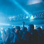 Intensyvūs balto stroboskopo šviesos spinduliai tamsioje renginio salėje virš šokančių žmonių.