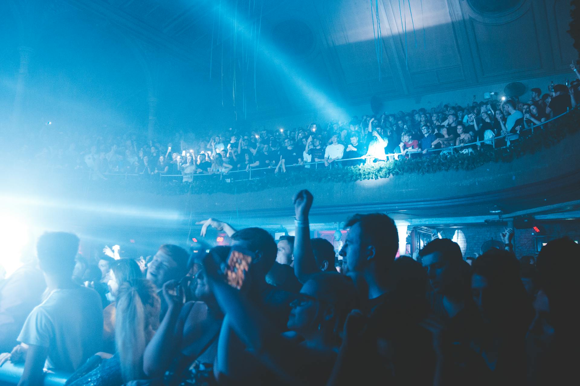 Intensyvūs balto stroboskopo šviesos spinduliai tamsioje renginio salėje virš šokančių žmonių.