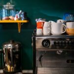 Modernus kavos aparatas stovi jaukioje namų virtuvėje šalia garuojančio kavos puodelio.